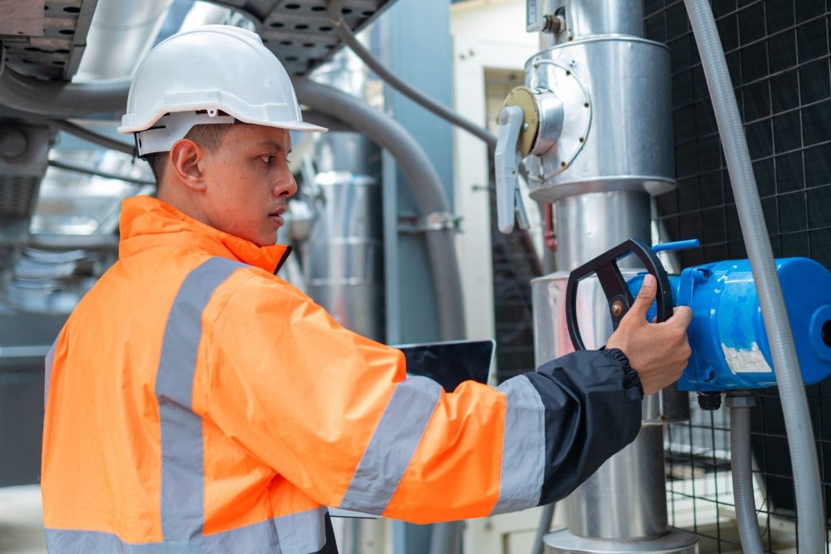 Worker in an orange safety jacket and helmet adjusting a blue water flow sensor in an industrial setting with pipes and machinery.